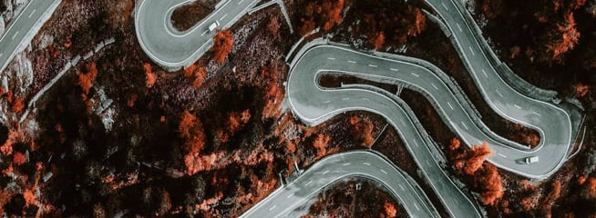 An aerial view of a winding road with multiple sharp curves and switchbacks, surrounded by autumn-colored trees.