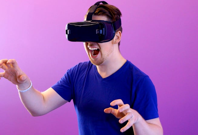 A person wearing a virtual reality headset and a blue T-shirt, standing against a purple background. The person has both hands raised and slightly curved, as if interacting with a virtual environment.